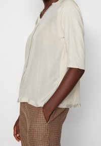 T-shirt de couleur crème à manches courtes avec un col en V, présentant une texture douce et une coupe légèrement ample. Associé à un pantalon brun à motifs.