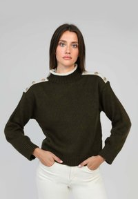 Pull en tricot vert foncé avec un col haut, présentant des accents crème et des boutons décoratifs sur les épaules. Manches et ourlet côtelés.