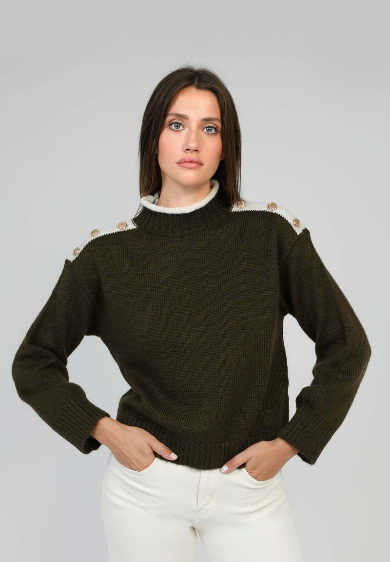 Pull en tricot vert foncé avec un col haut, présentant des accents crème et des boutons décoratifs sur les épaules. Manches et ourlet côtelés.
