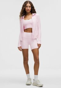 Conjunto deportivo rosa que incluye una sudadera con cremallera, una camiseta cropped y pantalones cortos. La tela es suave, con un diseño ajustado y sin patrones ni logotipos visibles.