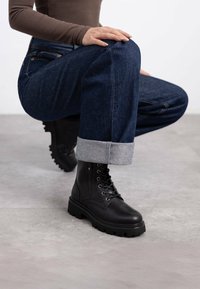 Botas de cuero negro con cordones y suela gruesa, con detalles de cremallera, combinadas con jeans de mezclilla oscuros con puños enrollados y una blusa ajustada marrón.