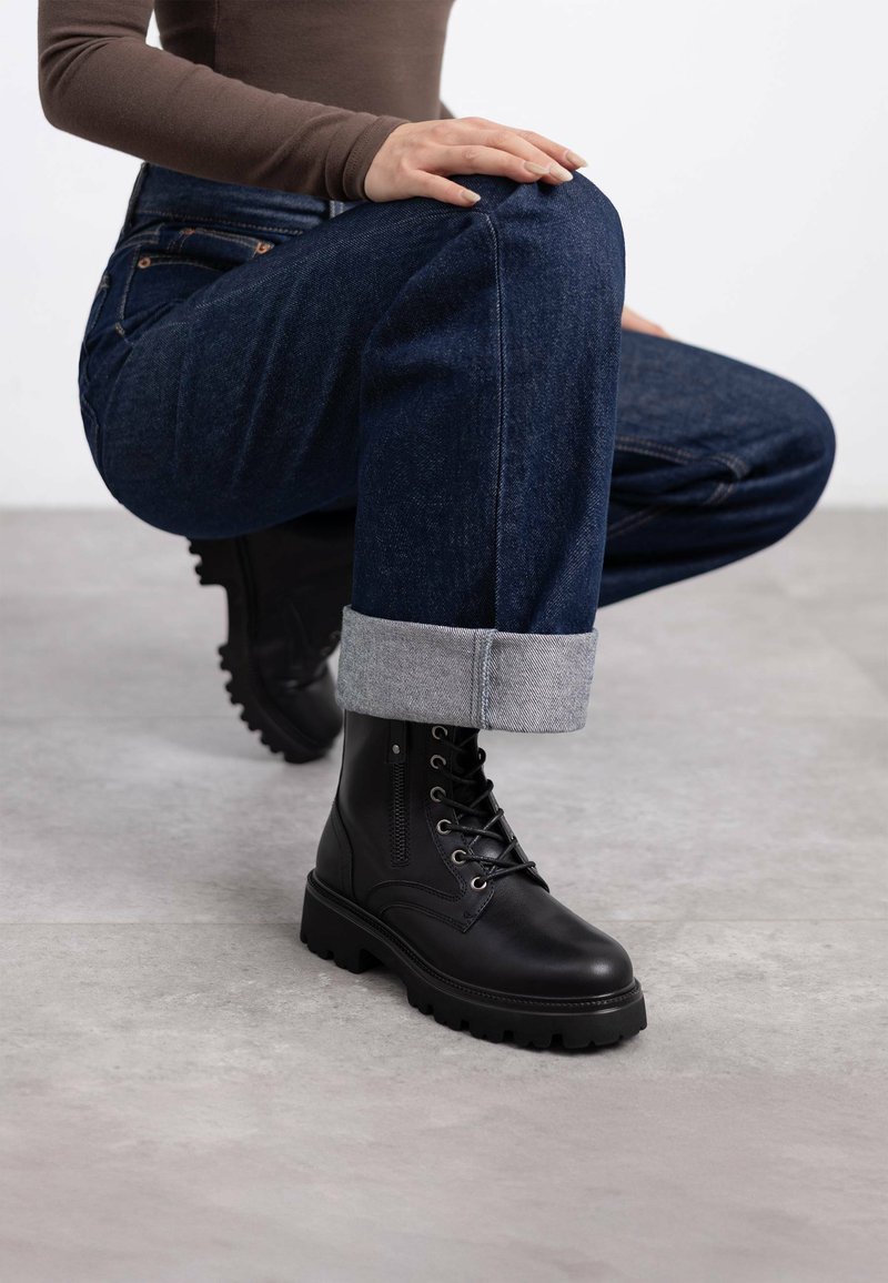 Botas de cuero negro con cordones y suela gruesa, con detalles de cremallera, combinadas con jeans de mezclilla oscuros con puños enrollados y una blusa ajustada marrón.