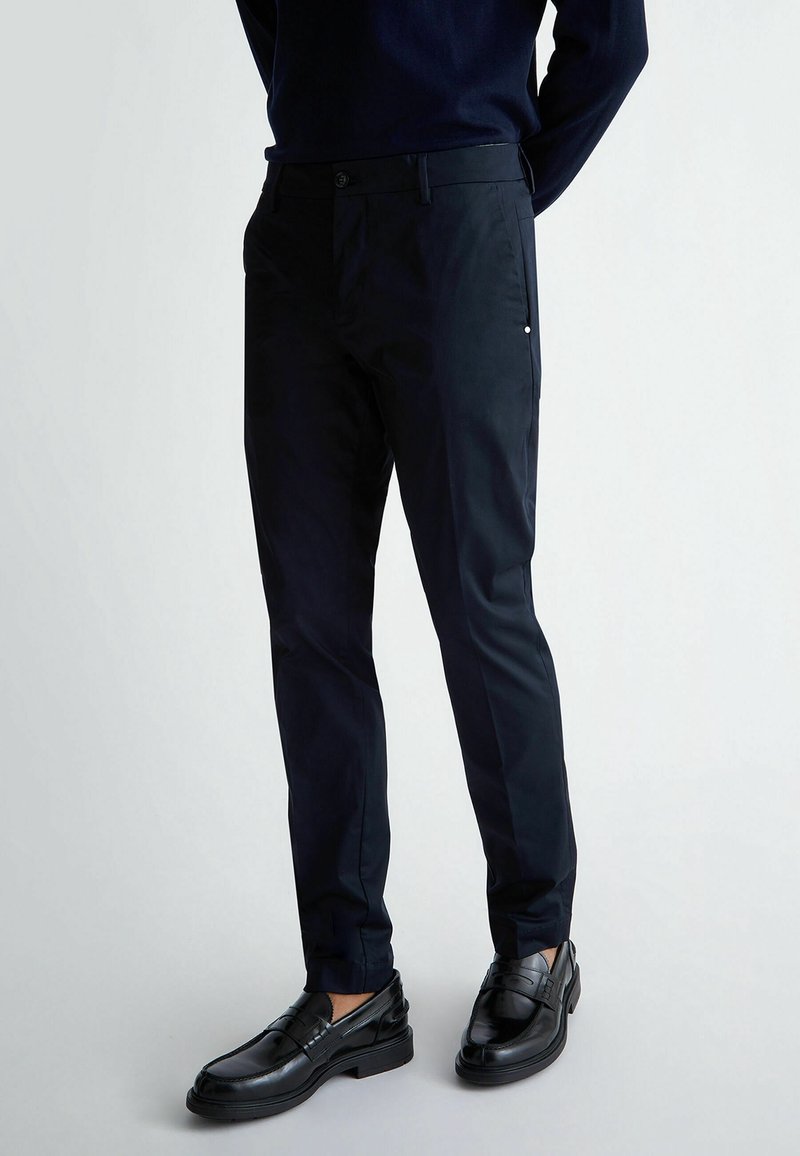 Homme portant un pantalon noir ajusté et des mocassins en cuir noir, debout devant un fond clair uni.