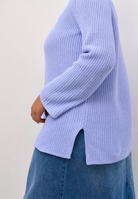 Pull en maille lavande claire avec un motif texturé, manches longues, fentes latérales et coupe ample, associé à une jupe en denim.