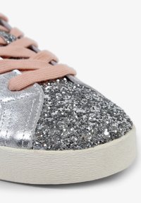 Zapatilla plateada brillante con una parte superior texturizada y brillante, con cordones rosas y una suela de goma blanca. La vista en primer plano captura los detalles.