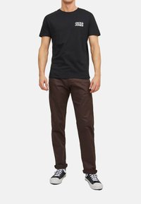 Black cotton t-shirt with white logo on left chest, paired with brown pants and black sneakers, featuring white laces and rubber soles.