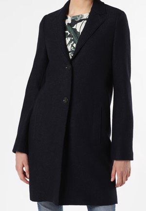 Femme portant un manteau en laine noir mi-long avec deux boutons sur le devant, sur un haut blanc imprimé de feuilles vertes.