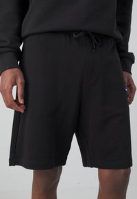 Shorts de sport noirs avec taille élastique à cordon de serrage, poches latérales et logo discret sur la cuisse gauche. Fabriqués en tissu doux et texturé.