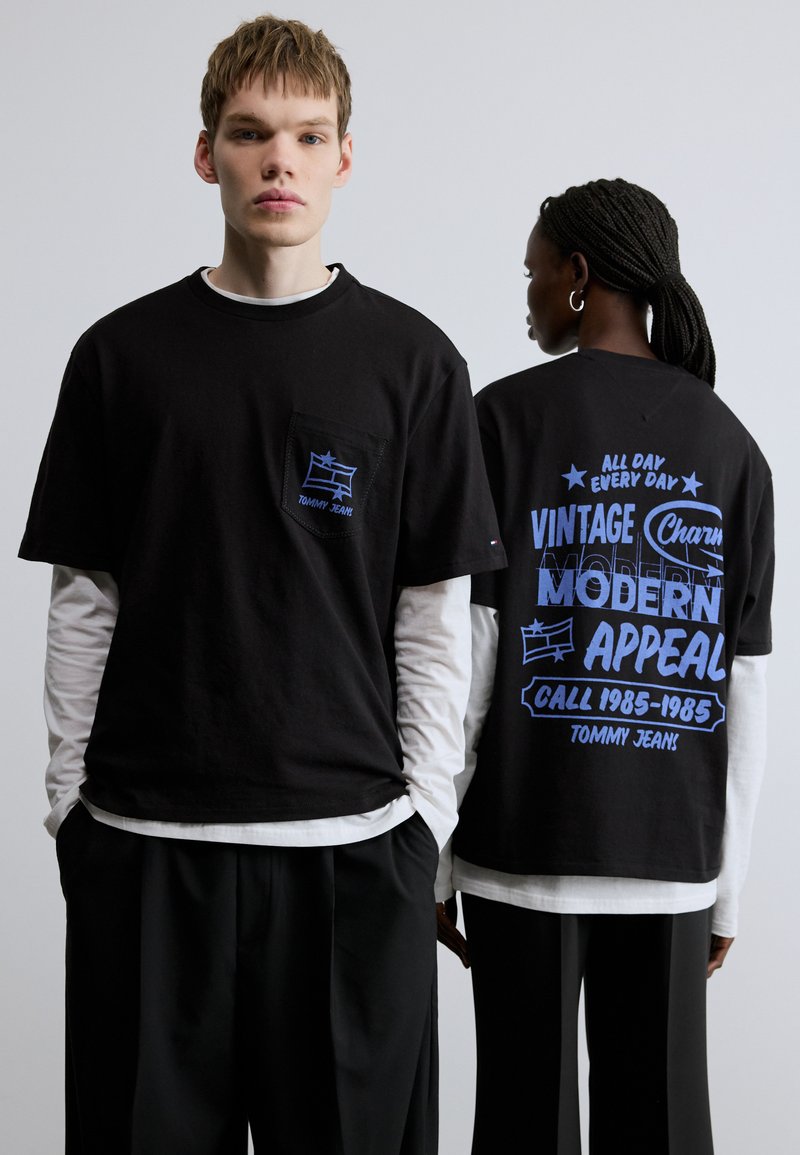 Čierne tričko s krátkym rukávom s modrými grafikami, ktoré má na hrudi vrecko a potlač na chrbte. Nosené cez biele tričko s dlhým rukávom. Voľný strih.