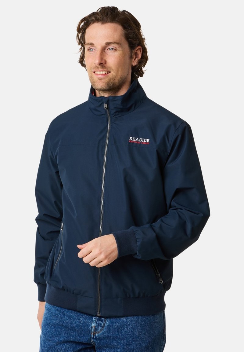 Mørkeblå letvægtsjakke med høj krave, frontlynlås og broderet "SEASIDE" logo. Har to sidelommer og strikkede manchetter.