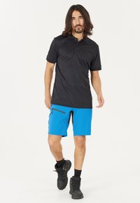 Svart polo-shirt med två knappar, parat med ljusblå shorts som har en dragkedjeficka och svarta detaljer. Modellen bär mörka vandringsskor.