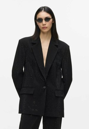 Femme aux cheveux noirs et raides portant de petites lunettes de soleil noires et un blazer noir clouté avec un pantalon assorti sur un fond blanc uni.