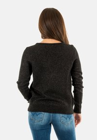 Pull noir avec un léger éclat, à manches longues et col rond. Porté avec un jean bleu avec des détails sur les poches arrière.