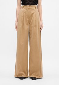 Beige bredde benbukser med høj talje, plisseret front og blød, glat struktur. Stoffet falder løst og har sidelommer.