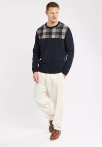 Homme portant un pull bleu marine avec un motif écossais beige, un pantalon crème et des chaussures en cuir marron à lacets, debout avec les mains dans les poches en regardant à gauche.