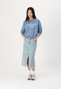 Blue sweatshirt with white logo, paired with a light denim skirt featuring a front slit. Model wears beige sneakers and white socks.