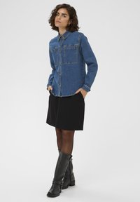 Denim jaka vidējā zilā krāsā ar pogu priekšpusi, divām krūšu kabatām un klasisku apkakli. Apvienota ar melnu svārku un melniem augstiem zābakiem.