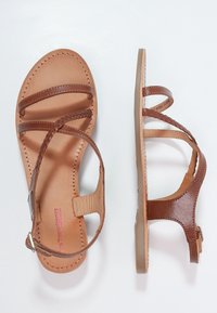 Hnědé kožené ploché sandály s pletenými a hladkými popruhy, s přezkou kolem kotníku a šitými okraji, zobrazené z horního a bočního pohledu na bílém pozadí.