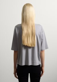 Persona con lunghi capelli biondi lisci, che indossa una maglietta grigio chiaro a maniche corte e pantaloni neri, di fronte a un muro bianco.