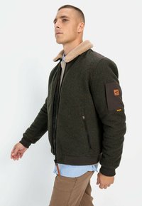Green bomber jacket with a shearling collar, brown textured sleeves, front zipper, and side pockets. Features a patch on the left arm.