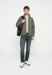 Chaqueta de plumas verde con cuello alto, suéter de punto beige, jeans oscuros y bolso bandolera negro. Calzado: zapatillas blancas con rayas negras.