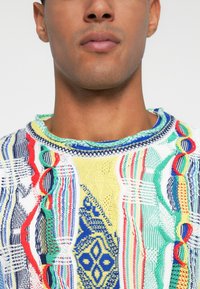 Χρωματιστό πλεκτό πουλόβερ με πολύπλοκα γεωμετρικά σχέδια σε μπλε, κόκκινο, πράσινο, κίτρινο και λευκό, με υφασματικές λεπτομέρειες και ευρύ λαιμό.