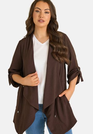 Kvinde med langt bølget hår iført en brun cardigan med åbent forstykke og opkørte ærmer over en hvid V-hals T-shirt og blå jeans, hænder rører ved cardiganen.