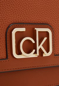 Borsa a mano in pelle sintetica marrone con una prominente accentuazione in oro del logo. Presenta cuciture in contrasto nere lungo i bordi.