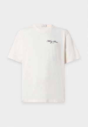 Jeune homme portant une chemise écrue à manches courtes avec un petit texte sur la poitrine, un pantalon gris délavé ample et des baskets noires et blanches, debout avec une main dans la poche.