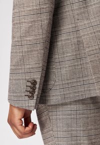 Braun karierter Blazerärmel mit einem strukturierten Stoff, vier dunklen Knöpfen am Manschetten, und einer taillierten Passform, die das Muster zur Geltung bringt.