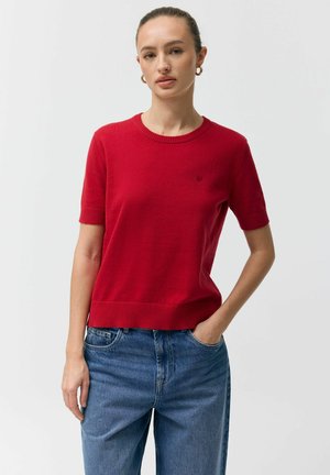 Kobieta z włosami związanymi do tyłu, nosząca złote kolczyki koła, czerwony krótki sweter z dzianiny oraz niebieskie dżinsy, z jedną ręką w kieszeni.