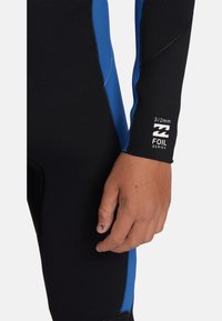 Muta nera con accenti blu, realizzata in neoprene 3/2 mm. La vista ravvicinata mostra le cuciture e il logo della serie nell'area del polso.