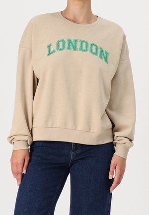 Beiger Pullover mit grünem "LONDON"-Schriftzug auf der Brust, getragen zu dunkelblauen Jeans an einer stehenden Person vor schlichtem Hintergrund.