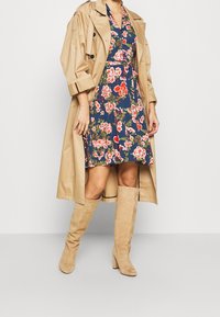 Kvinna iklädd en beige trenchcoat över en marinblå blommig klänning med knähöga beiga mockastövlar, stående mot en enkel bakgrund.