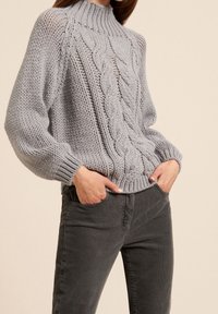 Femme portant un pull épais gris clair en maille torsadée à manches longues et un pantalon gris foncé, les mains dans les poches.