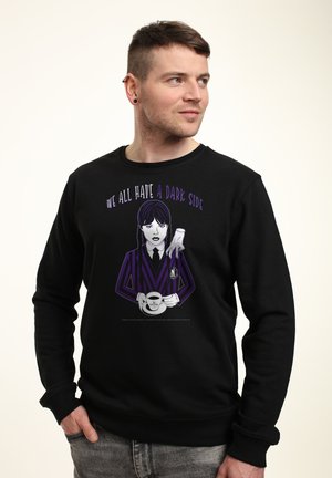 Mand iført sort sweatshirt med en lilla-stribet karakter, der holder en kaffekop, og teksten "Vi har alle en mørk side."