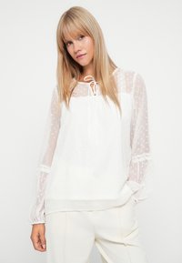 Blouse blanche en tissu transparent avec des pois, décolleté à cordon, et manches longues bouffantes avec des accents en dentelle. Texture lisse et légère.