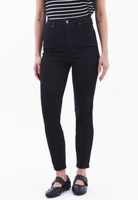 Jeans negros de talle alto hechos de denim, con un corte ajustado, diseño de cinco bolsillos y cierre con botón de metal. Llevados con zuecos negros.