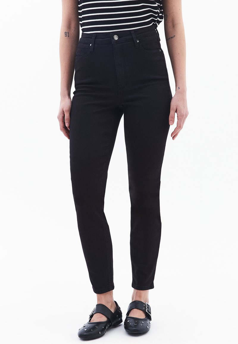 Jeans negros de talle alto hechos de denim, con un corte ajustado, diseño de cinco bolsillos y cierre con botón de metal. Llevados con zuecos negros.