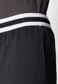 Close-up van zwarte sportshorts met een zwart-witte gestreepte elastische tailleband op een donkergrijze top.