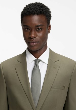 Unge mand med kort krøllet hår iført olivengrøn jakke, hvid skjorte og tekstureret gråt slips mod en ensfarvet hvid baggrund.