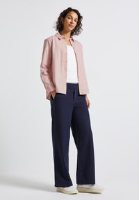 Donna in piedi, che indossa una camicia a righe rosa con bottoni sopra una maglietta bianca, pantaloni larghi blu scuro e sneakers bianche su uno sfondo bianco.