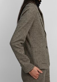 Brun och beige rutig kavaj med en figurnära design, två knappar och en enkel ficka. Materialet verkar texturerat och strukturerat.