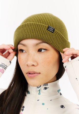 Olijfgroene geribbelde beanie met een zwart genaaid label, gedragen door een persoon; patroon en textuur zijn duidelijk met een opgerolde zoom.