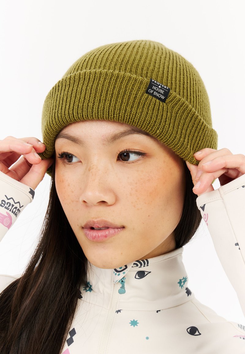 Olijfgroene geribbelde beanie met een zwart genaaid label, gedragen door een persoon; patroon en textuur zijn duidelijk met een opgerolde zoom.