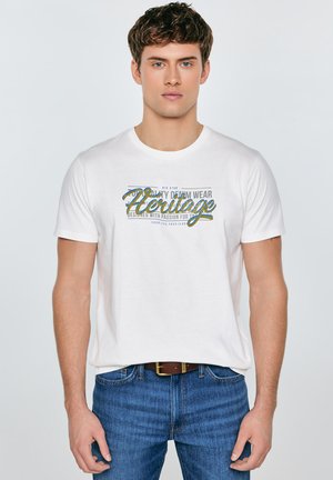 Biała bawełniana koszulka z kolorowym nadrukiem "Heritage". Modelka zestawia ją z niebieskimi jeansami i brązowym paskiem.