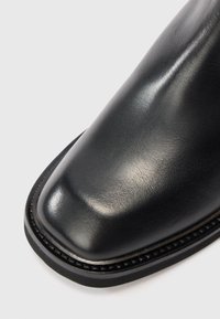 Chaussure en cuir noir avec bout carré et semelle cousue sur fond blanc, présentant une surface lisse et polie ainsi qu'une texture.