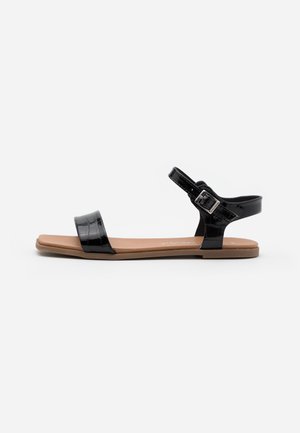 Sorte lakerede sandaler med flad sål, der har en bred fodrem og en justerbar ankelrem med en sølvspænde.