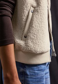 Crèmekleurige fleecevest met een ribbelzoom en zijzak, gecombineerd met een donkerbruine lange mouw top en lichtblauwe denim jeans.