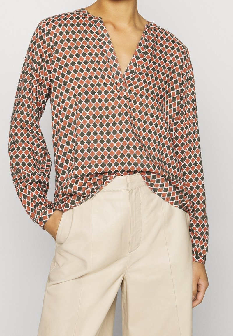 Blouse à manches longues dans un motif géométrique rouge brique et vert, présentant un décolleté en V et une coupe décontractée, associée à un pantalon en cuir beige.